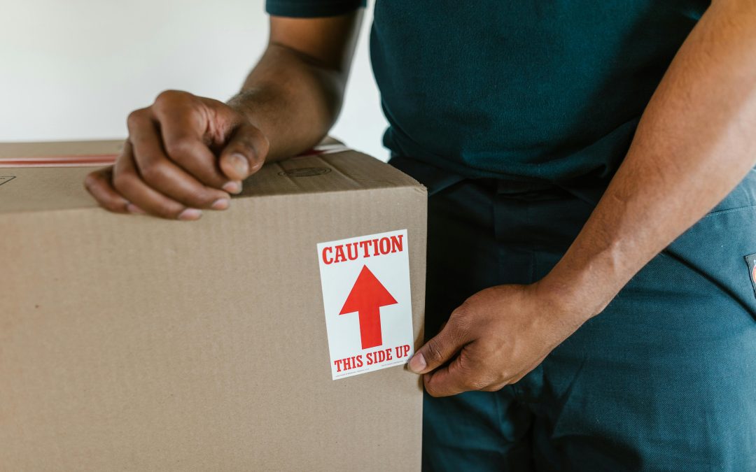 Persona sosteniendo una caja de cartón con una etiqueta que dice "CAUTION THIS SIDE UP" para prevenir daños en el transporte de muebles.